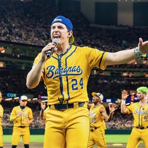 May include: A group of people in yellow baseball uniforms with blue detailing. The uniforms have the word "Bananas" and the number "24" on the front. The group is on a baseball field, with a crowd in the background.