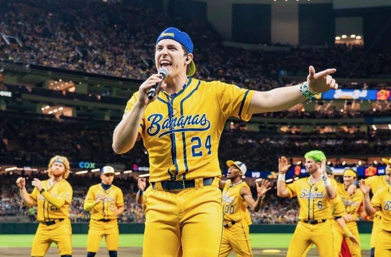 May include: A group of people in yellow baseball uniforms with blue detailing. The uniforms have the word "Bananas" and the number "24" on the front. The group is on a baseball field, with a crowd in the background.