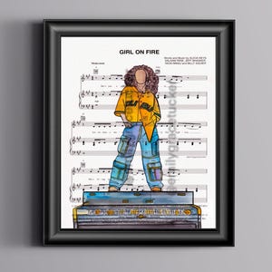 May include: Framed art print with a stylized illustration of a person on a piano. The person wears a yellow jacket and blue jeans. The background is sheet music, with the words "GIRL ON FIRE" at the top. The print is in a black frame.