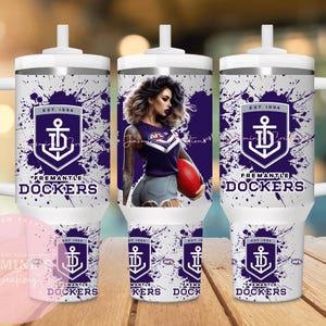 Puede incluir: Vaso blanco con asa, decorado con un diseño salpicado morado y blanco. Se muestra el logotipo y el texto de Fremantle Dockers. También aparece una mujer con una camiseta de Fremantle Dockers. El vaso tiene tapa y pajita blancas.