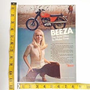 May include: Vintage magazine advertisement featuring a BSA motorcycle and a woman. The motorcycle is orange and black, with the text "BEEZA" and "The Bold Way To Make Time." The woman is wearing a light-colored top and brown pants.