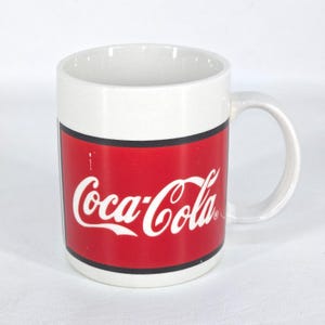 May include: White ceramic mug with a red band featuring the Coca-Cola logo in white script. The mug has a white handle and a white interior. A classic beverage accessory.
