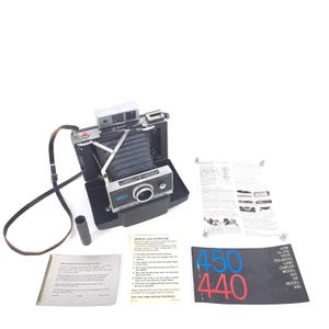 May include: A vintage Polaroid Land Camera Model 450 in a black case, with a silver and black body. Includes a flash attachment, lens, and a brown leather strap. Accompanying are instruction manuals and a guide with the numbers 450 and 440.