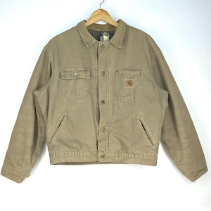 May include: A tan work jacket with a button-up front, two chest pockets, and two side pockets. The jacket has a brown collar and a small logo patch on the right chest pocket. It is hanging on a wooden hanger.