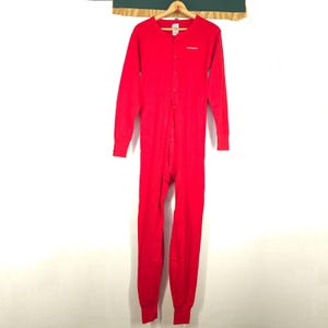 May include: A red, one-piece thermal jumpsuit with long sleeves and legs, hanging on a wooden hanger. The jumpsuit has a button-up front and the brand name "carhartt" is printed in white on the chest. The garment is made of a ribbed knit fabric.
