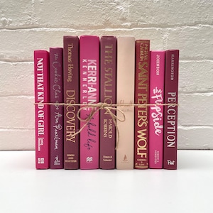 May include: A selection of books with pink and burgundy spines, bound with twine. Titles include "Not That Kind of Girl," "Discovery," and "Perception." The books are set against a white brick wall.