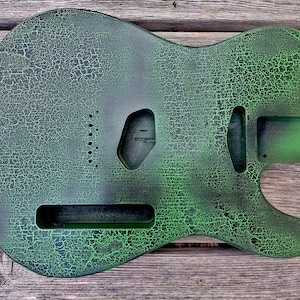 Könnte beinhalten: Ein Gitarrenkorpus mit einem verwitterten, rissigen Finish in Grün- und Schwarztönen. Der Korpus hat Aussparungen für Tonabnehmer, einen Hals und andere Hardware. Das Finish verleiht der Gitarre ein Vintage-Aussehen.