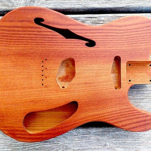 May include: A wooden guitar body with a reddish-brown finish. The body has a classic shape with a cutaway, f-hole, and various routed sections for pickups and hardware. The wood grain is visible.
