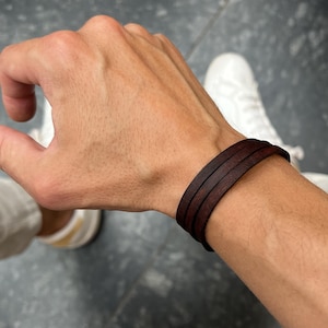 Könnte beinhalten: Ein dunkelbraunes Lederarmband mit drei gestapelten Bändern wird am Handgelenk getragen. Das Armband ist der Mittelpunkt, mit einem verschwommenen Hintergrund einer Hand und weißen Turnschuhen.