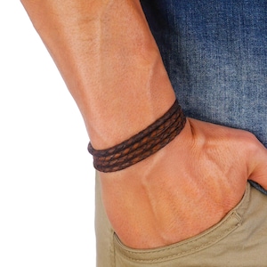 Può includere: Bracciale in pelle marrone intrecciata. Il bracciale è avvolto più volte attorno al polso. La pelle ha un aspetto strutturato e un colore marrone scuro. Il bracciale è indossato al polso di una persona.