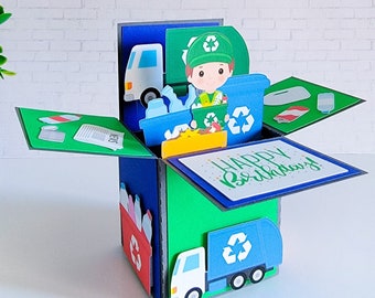 May include: A handmade pop-up birthday card featuring a recycling theme. The card is a box shape with green, blue, and red panels. It includes a recycling truck, a recycling bin, and a cartoon character. The words "Happy Birthday" are written on the front.