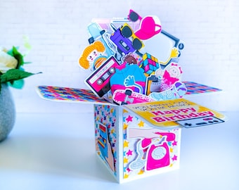 May include: A colorful pop-up card with a retro theme. The card features a variety of 80s and 90s icons, including a boombox, a Rubik's Cube, a cassette tape, a phone, and a skateboard. The card reads "You aren't old, you're retro! Happy Birthday".