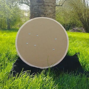 May include: The back of a round, brown cardboard shield with a white wooden frame. The shield has several small silver screws holding it together.
