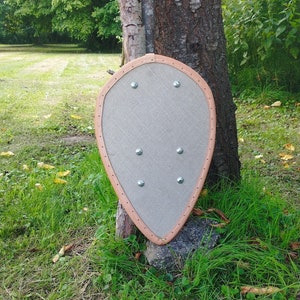 Peut inclure: Un bouclier en bois avec une bordure en cuir brun et des rivets métalliques. Le bouclier est appuyé contre un tronc d'arbre.