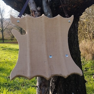 Puede incluir: Un objeto de madera marrón en forma de escudo con textura de arpillera y remaches metálicos. El escudo está colgado de una rama de árbol.