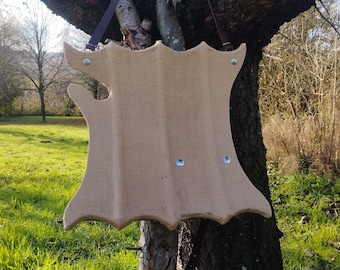 May include: A brown, wooden, shield-shaped object with a burlap texture and metal rivets. The shield is hanging from a tree branch.