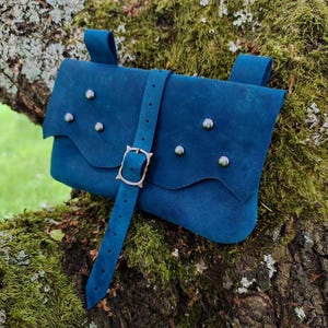 Può includere: Un borsello da cintura in pelle blu con chiusura a patta ondulata, un cinturino verticale con fibbia in metallo e sei borchie metalliche argentate disposte sul davanti. Il borsello presenta due passanti sul retro per l'aggancio alla cintura.