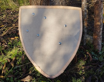May include: A medieval-style shield with a light tan fabric surface and a wooden frame. The shield has a rounded top and a pointed bottom. Six metal studs are evenly spaced across the fabric. The shield is displayed outdoors near trees and grass.