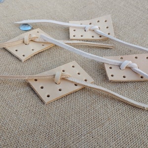May include: Four leather squares with holes punched in them. Each square has a white leather cord threaded through the holes and tied in a knot.