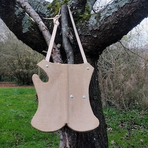 May include: A light brown leather armor with a strap hanging from a tree branch. The armor has a unique shape with a pointed bottom and a rounded top. There are four silver rivets on the front of the armor.