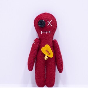 Könnte beinhalten: Handgefertigte rote Voodoo-Puppe mit weißen Nahtdetails. Die Puppe hat ein schwarzes Knopfauge, einen genähten Mund und einen gelben herzförmigen Aufnäher mit goldenen Nähten. Eine kleine goldene Glocke hängt am Herzen.