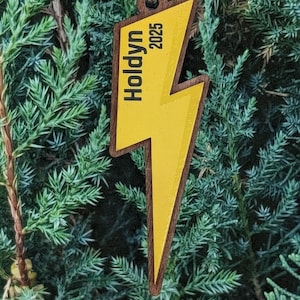 Könnte beinhalten: Holzschmuck in Form eines gelben Blitzes mit dem Namen "Holdyn" und dem Jahr "2025" in Schwarz aufgedruckt. Der Schmuck hängt vor einem Hintergrund aus grünem Laub.