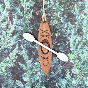 May include: Wooden kayak ornament with a white paddle. The brown ornament has the letters "XX" and "OK" etched into the wood. A natural twine loop is attached for hanging. The background is a backdrop of green foliage.