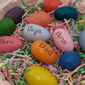 May include: A basket filled with colorful wooden eggs, each with a name written on them. The names include "Mom", "Dad", "Eliza", "Nora".