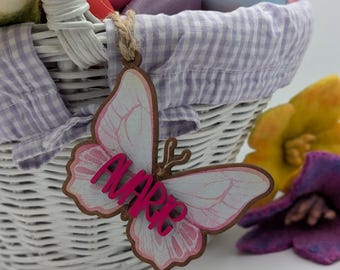 May include: A white wicker basket with a purple and white checkered liner is filled with colorful Easter eggs. A pink butterfly with the name "ALANA" written on it is hanging from the basket.