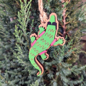 May include: A green wooden alligator ornament with pink and yellow details, wearing sunglasses and holding a skateboard. The ornament is hanging from a brown string.