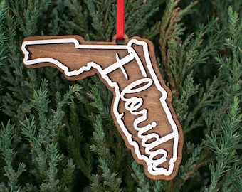 May include: Wooden Christmas ornament shaped like the state of Florida with the word "Florida" written in white inside the outline of the state. The ornament is hanging from a red ribbon.
