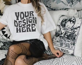 May include: A white t-shirt with the text "YOUR DESIGN HERE" in a retro font. The shirt is being worn by a person sitting on a bed with a black and white patterned throw pillow.