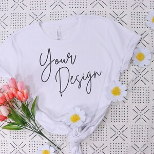 May include: White t-shirt with the text "Your Design" written in black cursive script. The shirt is lying on a white and black patterned surface with pink flowers and white daisies.