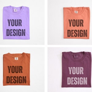 May include: Four folded t-shirts in different colors: lavender, coral, rust, and plum. Each shirt has the words "YOUR DESIGN" printed in a distressed font. The shirts are displayed on a white background.