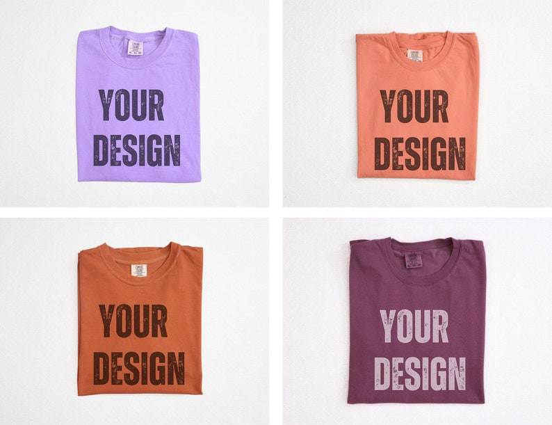 May include: Four folded t-shirts in different colors: lavender, coral, rust, and plum. Each shirt has the words "YOUR DESIGN" printed in a distressed font. The shirts are displayed on a white background.
