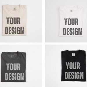 May include: Four folded t-shirts in cream, white, charcoal, and black. Each shirt has the words "YOUR DESIGN" printed in a distressed, gray font across the front. The shirts are displayed on a white background.