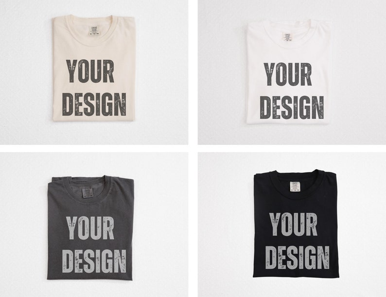 May include: Four folded t-shirts in cream, white, charcoal, and black. Each shirt has the words "YOUR DESIGN" printed in a distressed, gray font across the front. The shirts are displayed on a white background.