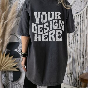 Puede incluir: Una camiseta gris oscuro de gran tama&ntilde;o con el texto "YOUR DESIGN HERE" impreso en blanco en la espalda. El texto est&aacute; en una fuente retro y ondulada.