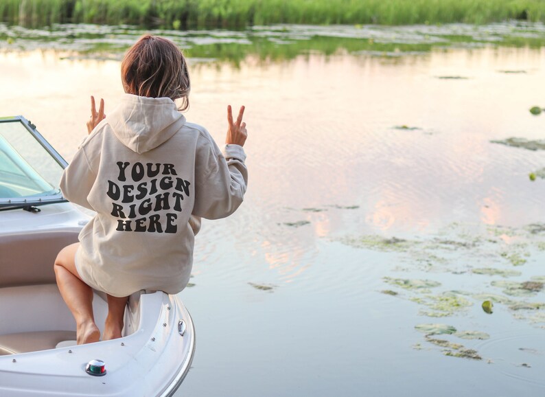 May include: A person wearing a beige hoodie with the text "YOUR DESIGN RIGHT HERE" on the back, sitting on a boat on a lake.