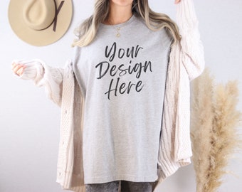 Può includere: Una maglietta grigio chiaro con la scritta "Your Design Here" in stile scritto a mano. La modella indossa un cardigan color crema. Un cappello marrone è appeso alla parete sullo sfondo.