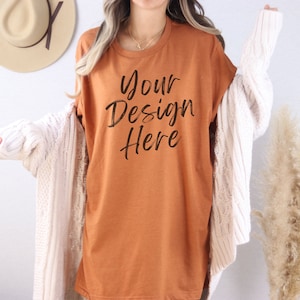 Puede incluir: Una camiseta de gran tamaño de color óxido con el texto "Your Design Here" impreso en la parte delantera. La camiseta la lleva puesta una persona con un cárdigan blanco.