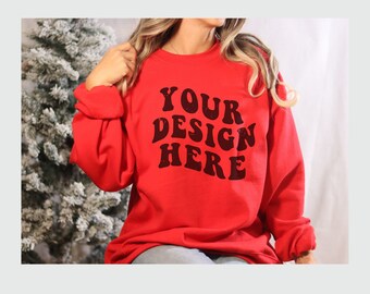May include: A red sweatshirt featuring the text "YOUR DESIGN HERE" in a retro font. The sweatshirt has long sleeves and a crew neck. The image is set against a white backdrop with a frosted evergreen tree in the background.
