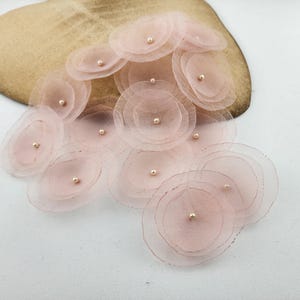 May include: A collection of blush pink layered fabric flowers with a pearl center. The flowers are arranged in a cluster on a light brown surface.