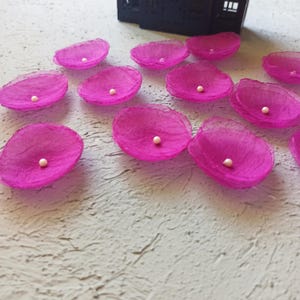May include: Magenta fabric petals, each with a small white bead centre. The petals are arranged on a textured, light grey surface. A black object is in the background. The petals appear to be handmade.