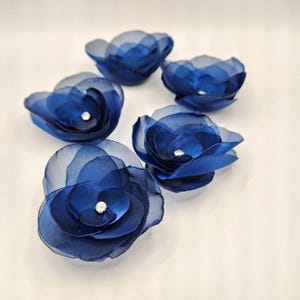 Könnte beinhalten: Fünf marineblaue Stoffblumen mit einem geschichteten Blütenblattdesign und einem kleinen, funkelnden Strassstein in der Mitte. Die Blumen sind auf einer weißen Oberfläche angeordnet und zeigen ihre zarte, durchscheinende Textur. Geeignet für Bastelarbeiten.