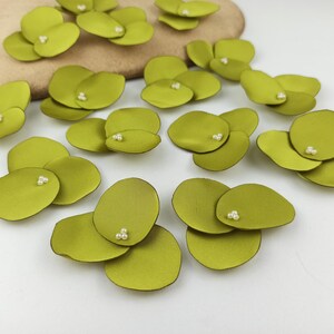 May include: A collection of green fabric flower petals with pearl accents. The petals are arranged on a light brown surface.
