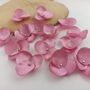 May include: Pink satin flower petals with a small rhinestone in the center. The petals are arranged on a white surface with a tan, heart-shaped object in the background. These are ideal for craft projects.