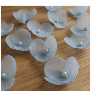 Könnte beinhalten: Hellblaue Stoffblumen-Verzierungen mit einer Perlenperle in der Mitte. Die durchscheinenden Blütenblätter sind in einer floralen Form angeordnet. Diese Bastelmaterialien sind auf einer Holzoberfläche verstreut.