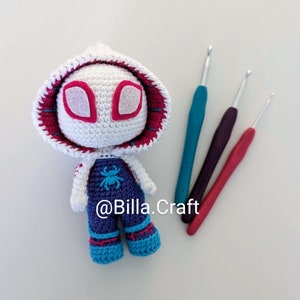 Może przedstawiać: Biała pluszowa zabawka amigurumi wykonana na szydełku z różowym wzorem pająka na twarzy, ubrana w niebieskie ogrodniczki z grafiką pająka i niebieskie buty. Zabawka leży na białym tle z trzema szydełkami.