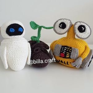 Puede incluir: Juguetes de peluche tejidos a crochet de WALL-E y EVE de la película animada WALL-E. WALL-E es amarillo con un diseño rojo y negro en su pecho. EVE es blanca con ojos azules. Una pequeña planta verde está entre ellos.